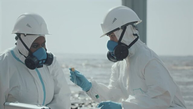 Two female ecologists wearing protective coveralls and respiratory masks examining test tubes with samples of polluted water and talking while working at gulf coast