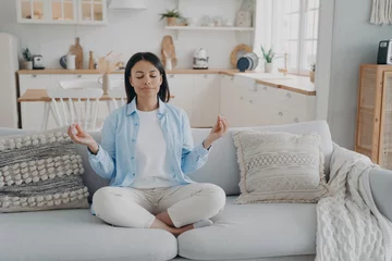 Tuinposter Serene woman practices yoga, meditates on couch in lotus position. Breathing exercises for mental balance, anxiety relief. Healthy lifestyle at home. © VK Studio