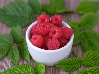 Red raspberries in a white bowl surrounded by raspberry leaves