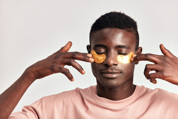 Young handsome black guy applying cosmetic under eye hydrogel patches on his face. African American millennial man practices skin care routine to keep healthy and youthful looking. Studio portrait.