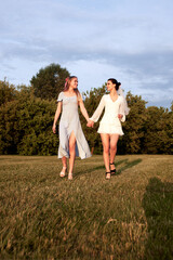 Charming bride in a white dress with white veil and a bridesmaid in a blue dress are walking in a summer park, holding hands, smiling happily, looking at each other. Cheerful bachelorette party.