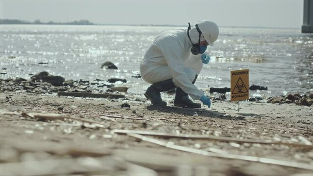 Ecologist wearing protective coverall and respiratory mask collecting sample of soil on polluted gulf shore with warning biohazard sign