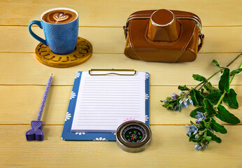 Old camera, blue notebook, cup of cappuccino, compass, exotic flower on yellow background.  Vacation plans concept