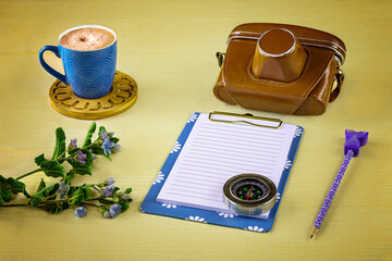 Old camera, blue notebook, cup of cappuccino, compass, exotic flower on yellow background.  Vacation plans concept