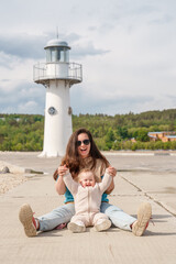 Fototapeta na wymiar Mom and daughter - a little girl baby toddler - sit on the background of a lighthouse in summer