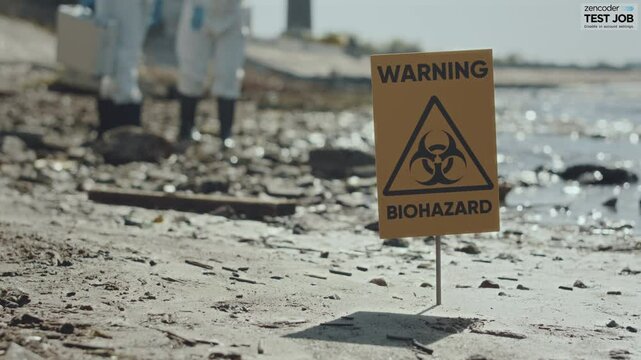 Close up selective focus shot of yellow warning biohazard sign on lakeshore in polluted area, group on ecologists working in background