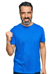 Middle aged man with beard wearing casual blue t shirt angry and mad raising fist frustrated and furious while shouting with anger. rage and aggressive concept.