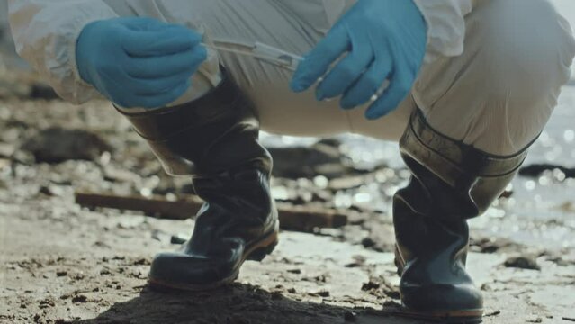 Ecologist wearing protective coverall, rubber boots and respiratory mask crouching on polluted shore, collecting sample of soil and examining test tube