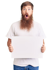 Handsome young red head man with long beard holding blank empty banner scared and amazed with open...