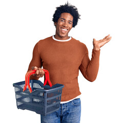 Handsome african american man with afro hair holding supermarket shopping basket celebrating...