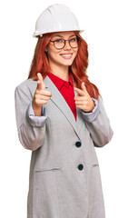 Young redhead woman wearing architect hardhat pointing fingers to camera with happy and funny face. good energy and vibes.