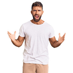 Young hispanic man wearing casual white tshirt crazy and mad shouting and yelling with aggressive expression and arms raised. frustration concept.