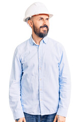 Young handsome man wearing architect hardhat looking away to side with smile on face, natural expression. laughing confident.