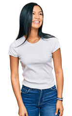 Beautiful hispanic woman wearing casual white tshirt looking away to side with smile on face,...