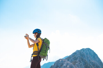 Fototapeta na wymiar climber on top of the mountain takes a photo. Girl with a backpack and hard hat in the mountains. Woman traveler takes a photo on a smartphone, takes pictures of beautiful mountain scenery.