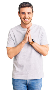 Handsome young man with bear wearing casual tshirt praying with hands together asking for forgiveness smiling confident.