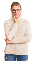 Young beautiful blonde woman wearing turtleneck sweater and glasses looking confident at the camera smiling with crossed arms and hand raised on chin. thinking positive.