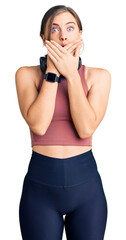 Beautiful caucasian young woman wearing gym clothes and using headphones shocked covering mouth with hands for mistake. secret concept.