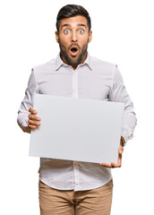 Handsome hispanic man holding blank empty banner scared and amazed with open mouth for surprise,...