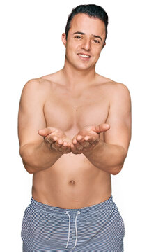 Handsome young man wearing swimwear shirtless smiling with hands palms together receiving or giving gesture. hold and protection