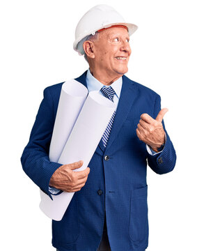 Senior handsome grey-haired man wearing architect hardhat holding blueprints pointing thumb up to the side smiling happy with open mouth