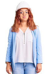 Young latin woman wearing architect hardhat puffing cheeks with funny face. mouth inflated with air, crazy expression.