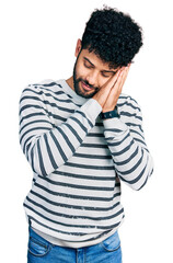 Young arab man with beard wearing casual striped sweater sleeping tired dreaming and posing with hands together while smiling with closed eyes.