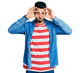 Young hispanic man with beard wearing casual denim jacket trying to open eyes with fingers, sleepy...