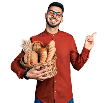 Young hispanic man with beard holding wicker basket with bread smiling happy pointing with hand and finger to the side