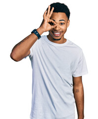 Fototapeta premium Young african american man wearing casual white t shirt doing ok gesture with hand smiling, eye looking through fingers with happy face.