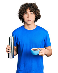 Caucasian teenager holding cup of coffee and thermo puffing cheeks with funny face. mouth inflated with air, catching air. - obrazy, fototapety, plakaty