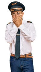 Handsome man with beard wearing airplane pilot uniform shocked covering mouth with hands for...