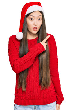 Young chinese woman wearing christmas hat surprised pointing with finger to the side, open mouth amazed expression.