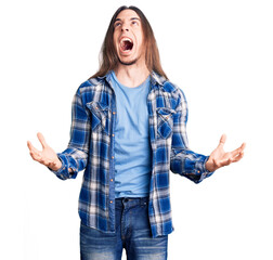 Young adult man with long hair wearing casual shirt crazy and mad shouting and yelling with aggressive expression and arms raised. frustration concept.