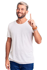 Young handsome blond man wearing casual t-shirt showing and pointing up with finger number one while smiling confident and happy.