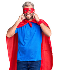 Young blond man wearing super hero custome covering ears with fingers with annoyed expression for...