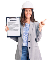 Young beautiful brunette woman wearing architect hardhat holding contract smiling happy pointing with hand and finger to the side