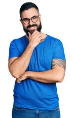 Hispanic man with beard wearing casual t shirt and glasses looking confident at the camera smiling with crossed arms and hand raised on chin. thinking positive.