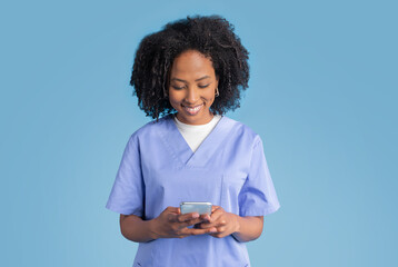 Positive young black lady doctor nurse in blue uniform typing on smartphone, chatting with client