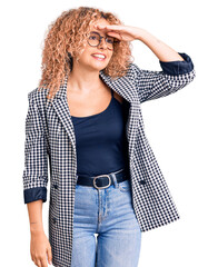 Young blonde woman with curly hair wearing business jacket and glasses very happy and smiling looking far away with hand over head. searching concept.