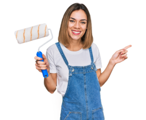 Young blonde girl holding roller painter smiling happy pointing with hand and finger to the side