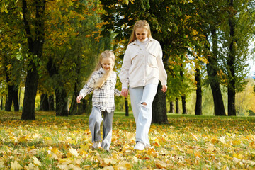 Naklejka na ściany i meble Family spend time together in autumn park. Mother and daughter are walking in the autumn park and throwing leaves up with their feet
