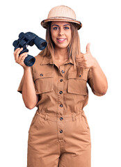 Young beautiful woman wearing explorer hat holding binoculars smiling happy and positive, thumb up doing excellent and approval sign