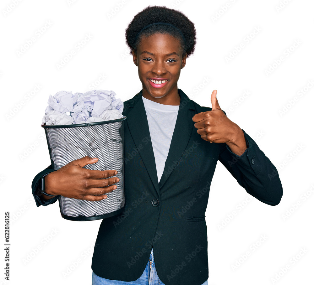 Sticker Young african american girl holding paper bin full of crumpled papers smiling happy and positive, thumb up doing excellent and approval sign