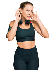 Obraz na płótnie Canvas Beautiful blonde woman wearing gym clothes and using headphones trying to hear both hands on ear gesture, curious for gossip. hearing problem, deaf