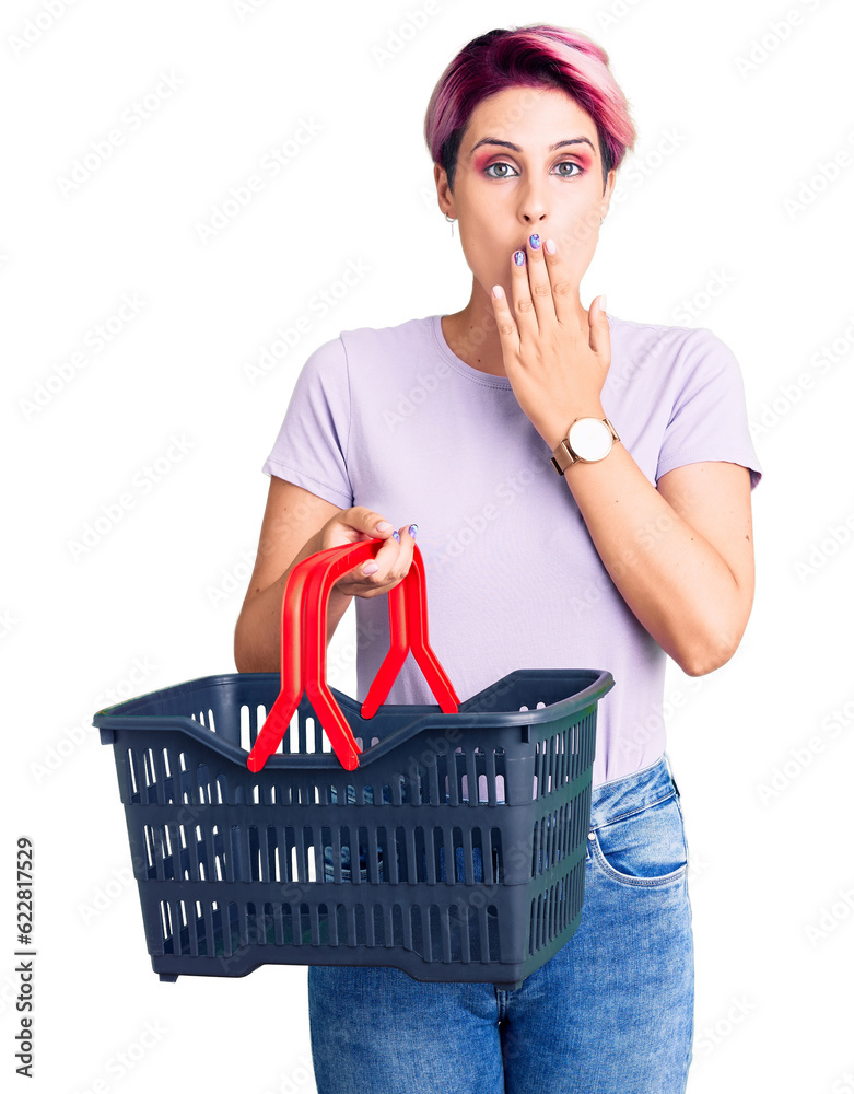 Sticker young beautiful woman with pink hair holding supermarket shopping basket covering mouth with hand, s