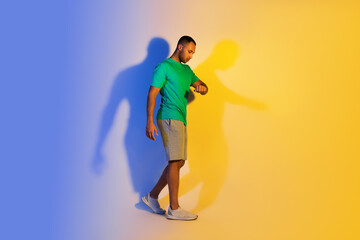 Fototapeta na wymiar Black Fitness Guy Looking At Smartwatch, Blue And Yellow Background