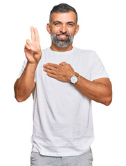 Middle age handsome man wearing casual white tshirt smiling swearing with hand on chest and fingers...