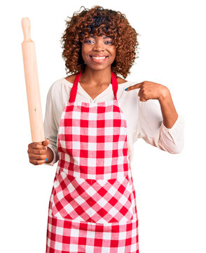 Young african american woman wearing apron holding rolling pin pointing finger to one self smiling happy and proud