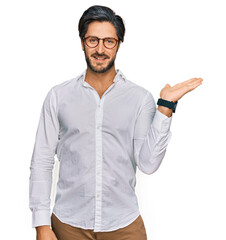 Young hispanic man wearing business shirt and glasses smiling cheerful presenting and pointing with palm of hand looking at the camera.